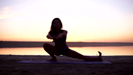 a sports brunette doing stretching exercises on a mat on the beach or a lake. accelerated footage. front view