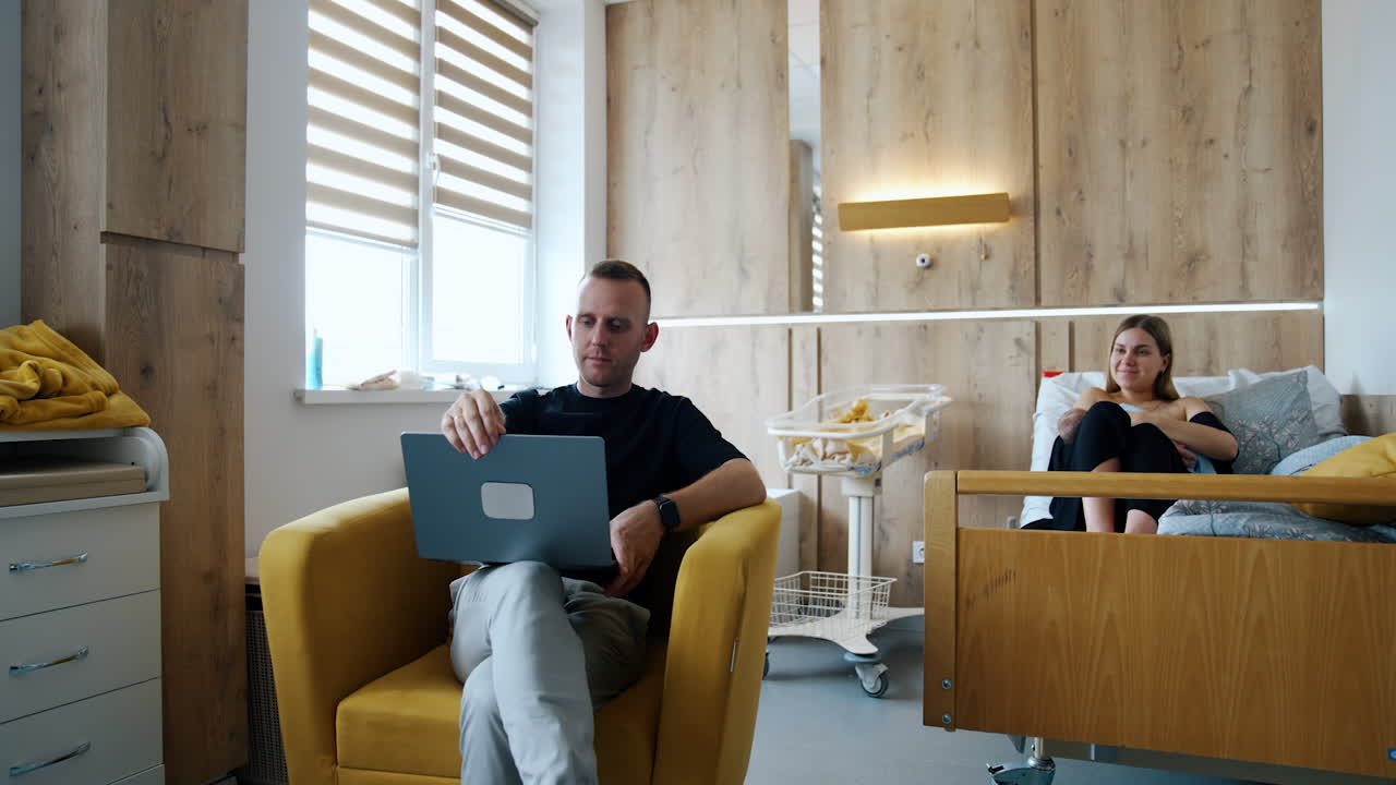 New-made Caucasian parents are in the hospital ward after childbirth. Man sits to the armchair with computer and woman breastfeeding her child.