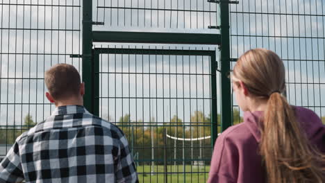 back view of youngsters entering volleyball court as coach opens green gate, with a distant football field and lush trees visible in the background