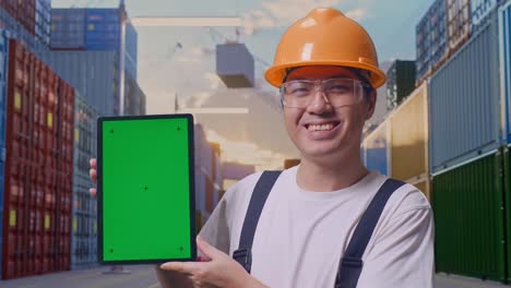 Close Up Of Asian Man Worker Wearing Goggles And Safety Helmet Smiling And Showing Green Screen Tablet To The Camera While Standing At Container Yard Warehouse