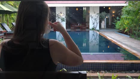 Over-the-Shoulder-Shot-of-a-Young-Woman-Sipping-Tea-While-Looking-at-the-Hotel-Swimming-Pool