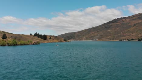 Cámara-Lenta---Antena---Barco-En-El-Lago-Dunstan,-Otago-Central,-Nueva-Zelanda-Con-Montañas-Y-Nubes-En-El-Fondo