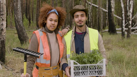 african american woman activist and caucasian male coworker smiling at the camera while they holding a rake and small trees