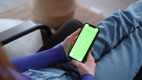 young woman sitting on sofa at home and uses smartphone with green screen mock-up. holding in hand and looking on the screen. 4k