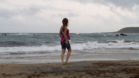 Cinematic-shot-at-sunrise-of-a-woman-walking-along-the-shore-of-Las-Canteras-beach-and-where-the-waves-break-on-the-shore