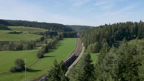 Drone-shot-showcasing-a-red-train-with-multiple-carriages,-coursing-on-a-single-track-railway-line-through-a-green-field,-with-a-backdrop-of-trees-under-a-clear-blue-sky