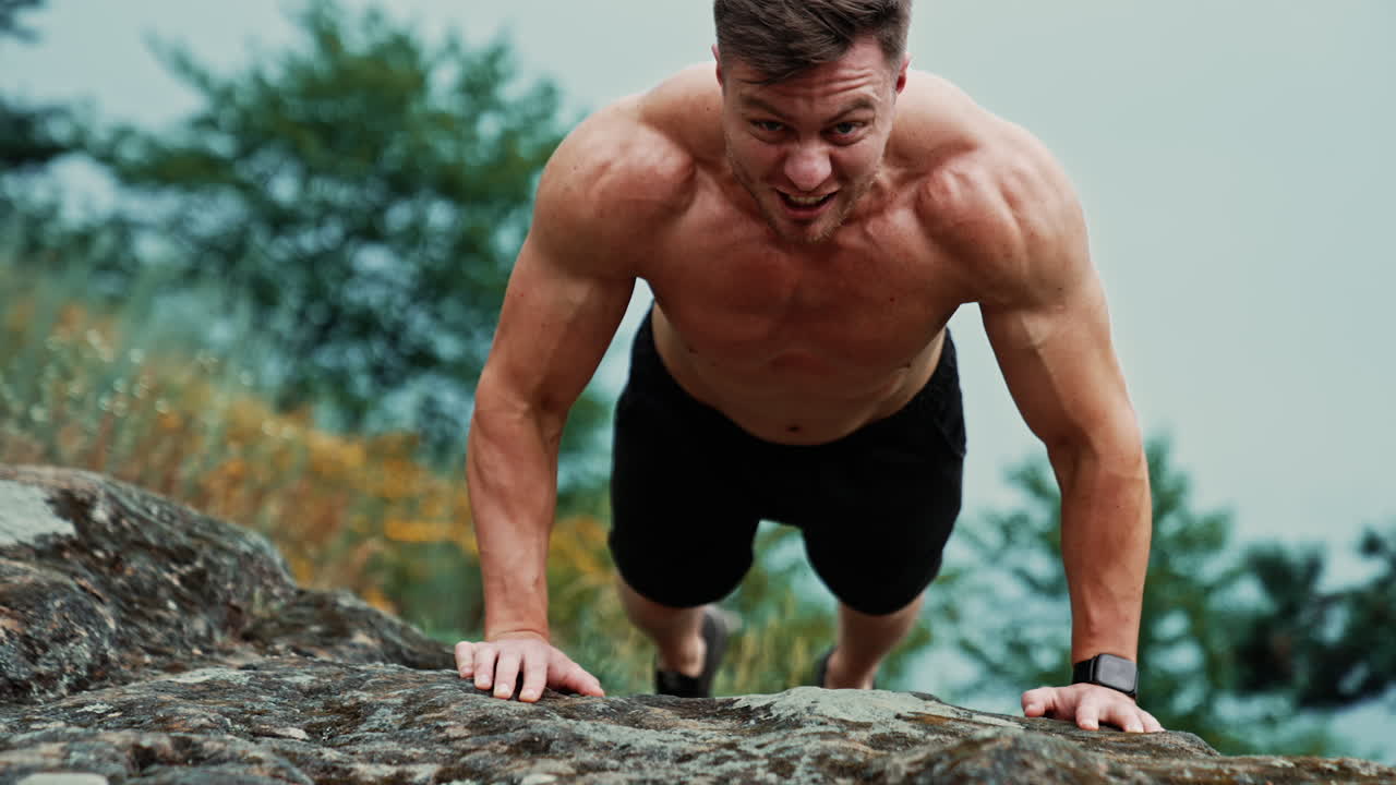 Sportsman smiles doing push-ups. Strong man working out on the rock looking at camera.
