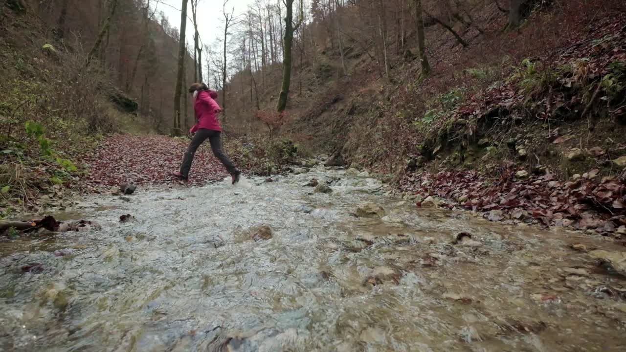 Lady crossing a small river on a walking path in slow motion. Jumping over the water in hiking boots and hiking clothes during a dull day. Pokljuka Gorge in Slovenia Triglav National Park