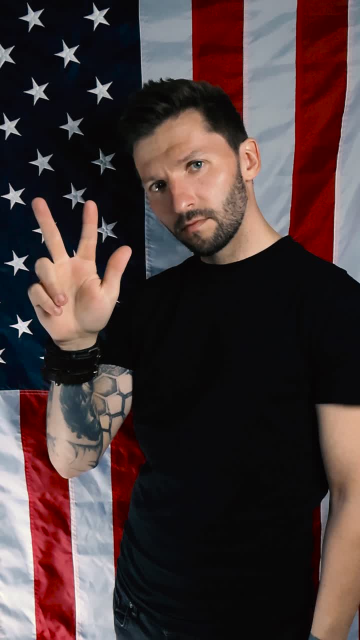 Tattooed man countring to three with his arm raised and using his fingers in slow motion in front of a vertical United States of America or american flag. Man is determined and focused. One Two Three