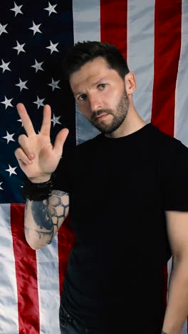 tattooed man countring to three with his arm raised and using his fingers in slow motion in front of a vertical united states of america or american flag. man is determined and focused. one two three