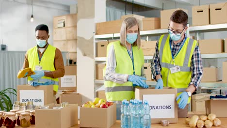 multiethnic group of senior and young volunteers in facial masks packing food and clothes in donation boxes in charity warehouse