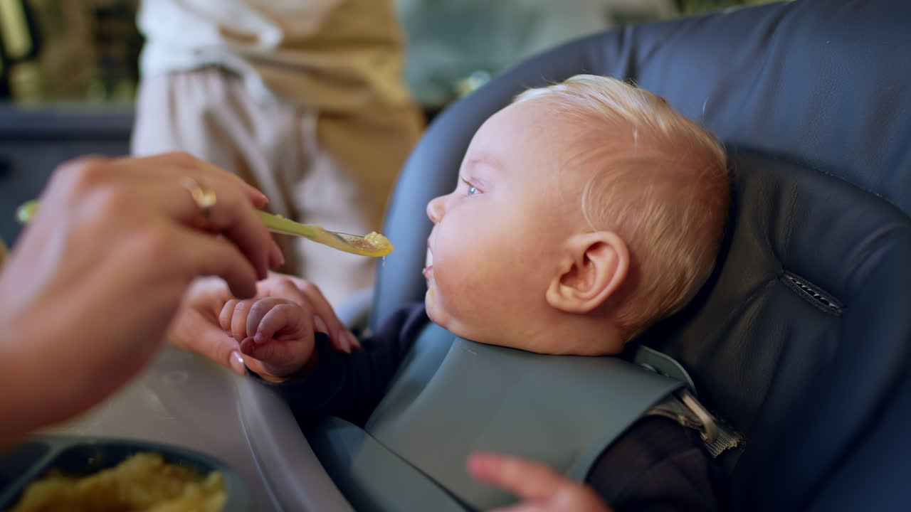 Chubby little infant starting to cry at the high chair. Mom gives a spoon with food to her child. Close up.