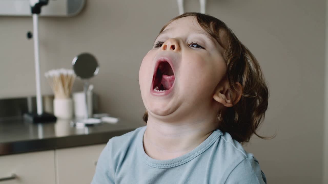 Bambino che apre la bocca con una maglietta blu, si inclina, mostra la gola e chiude con un sorriso compiaciuto in una clinica con specchio