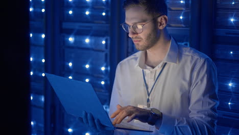 Male network engineer doing a system check standing in the server room with his laptop. At data center men server specialists inspecting working system and hardware of rack server computer cabinets