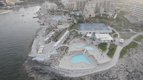 Aerial-view-of-a-seaside-resort-with-multiple-pools,-a-tennis-court,-and-moored-boats,-nestled-between-a-rocky-shore-and-upscale-buildings