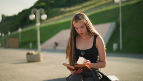 mujer sentada al aire libre en un banco con las piernas cruzadas, volviendo una página de su libro y observando pensativamente un pedazo de papel, luego dejándolo caer de nuevo, con rasgos de fondo borrosos postes de lámparas