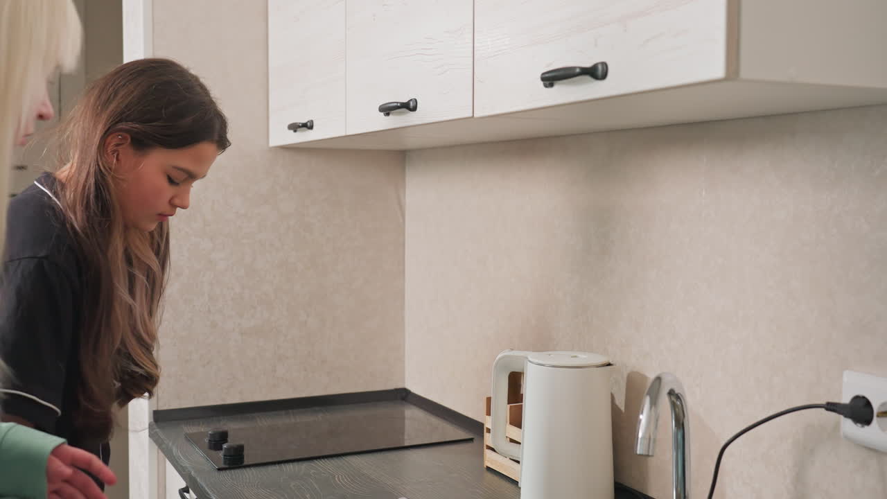 woman opening kitchen cabinet to collect knife and plate from upper cabinets while friend gives her something kitchen preparation cooking scene showing interaction and utensils modern