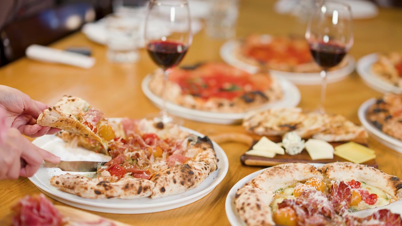 Primo piano di una mano che taglia pizza fresca su un tavolo di legno con vino e tagliere di formaggi