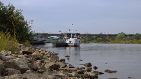 small travel ferry boat docking to pier on elbe river in pirna, germany with elbe bridge in background - riverbank timelapse