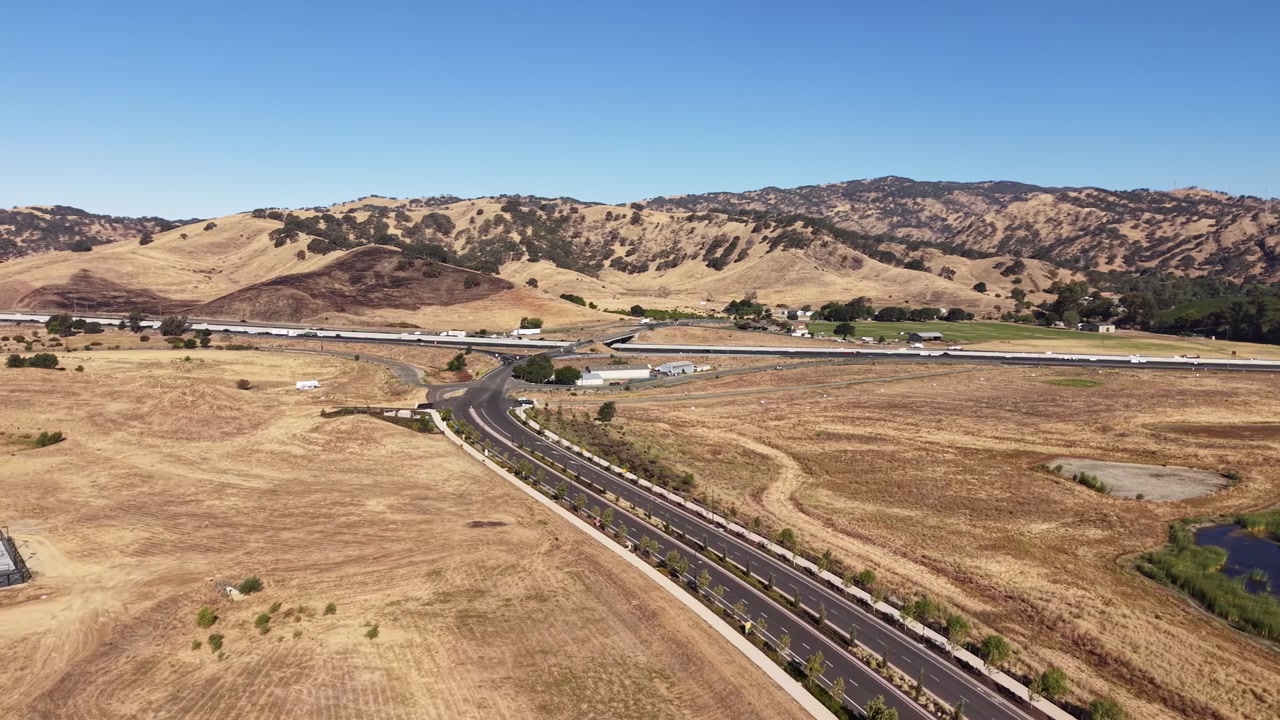 Scenic aerial view of the arid, golden-brown landscape of Lagoon Valley Park in Vacaville, California, showing a highway with traffic passing through the dry, rolling hills on a sunny day