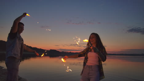 young happy multiethnic couple holding burning sparkling candles and running by the sea during sunset. slow motion shot