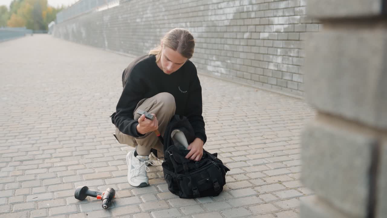 White photographer crouching by backpack remote, checking drone controller and gimbal, compact camera gear on pavement, brick wall backdrop, focused setup for shoot, content creation energy