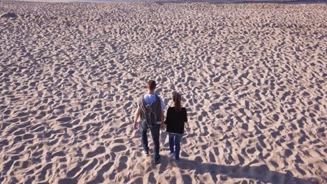 Toma-Aérea-De-Una-Pareja-Joven-Caminando-En-La-Playa-De-Arena,-Tomándose-De-La-Mano-Y-Corriendo-Hacia-El-Agua