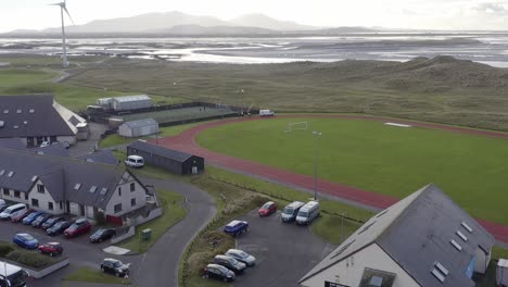 Tilting-drone-shot-of-the-track-and-pitch-in-Benbecula,-as-well-as-the-nearby-wind-turbine,-beach,-sea-and-mountains