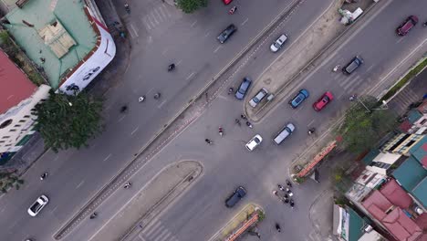 This-aerial-footage-captures-a-dynamic-intersection-in-Hanoi,-Vietnam,-showcasing-the-bustling-traffic-flow-of-cars,-motorcycles,-and-pedestrians