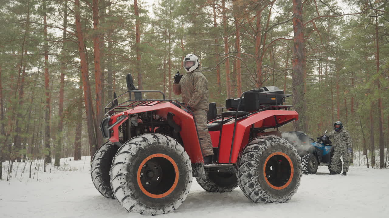 ATV rosso in una foresta di pini innevata, parcheggiato con piloti con casco nelle vicinanze, pneumatici tassellati sovradimensionati bordati di neve, fredda luce invernale che filtra attraverso i pini alti, pilota che esegue un controllo pre-corsa, robusto.