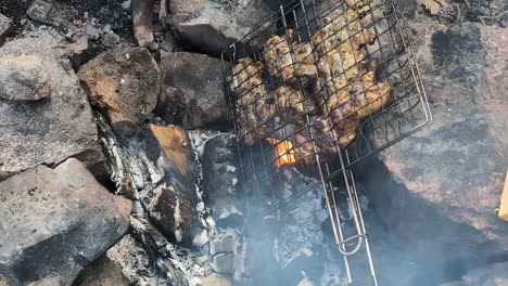 Meat-on-a-grill-to-bbq-on-the-beach-in-the-sand,-fireplace-with-rocks-around,-closeup-in-slow-motion,-static-shot