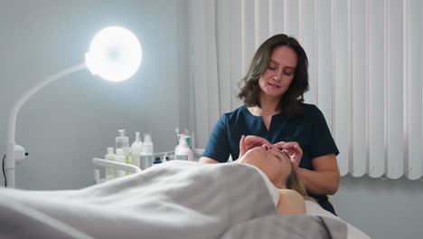 skin therapist at station smiling while performing gentle fingertip facial detox on relaxed client lying under blanket beside ring light and skin care tools on trolley in bright treatment room