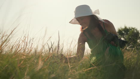 woman in green dress and wide brim hat walks through tall grass holding camera, pauses near flower and bends slightly to observe it with calm expression under soft afternoon sunlight in nature field