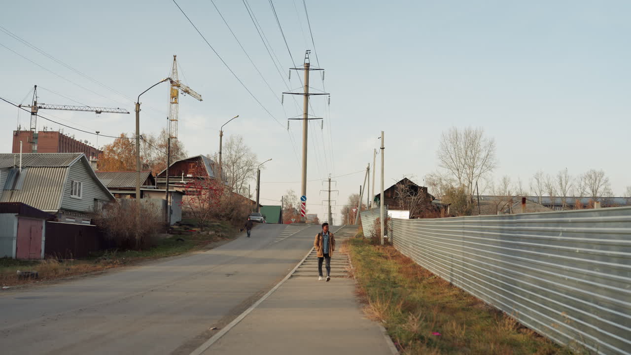 White man walking rural road repair, corrugated metal fence at right, wooden houses and shed at left, power poles and crane in distance, autumn light and calm atmosphere, solitary maintenance worker