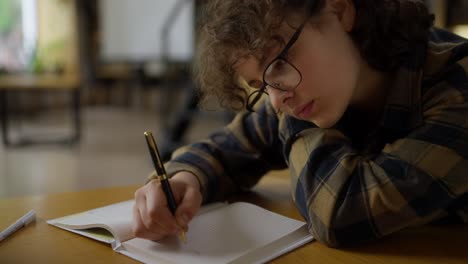 a tired teenage girl with curly hair in a checkered shirt and glasses makes notes in a book while sitting at a table in the library