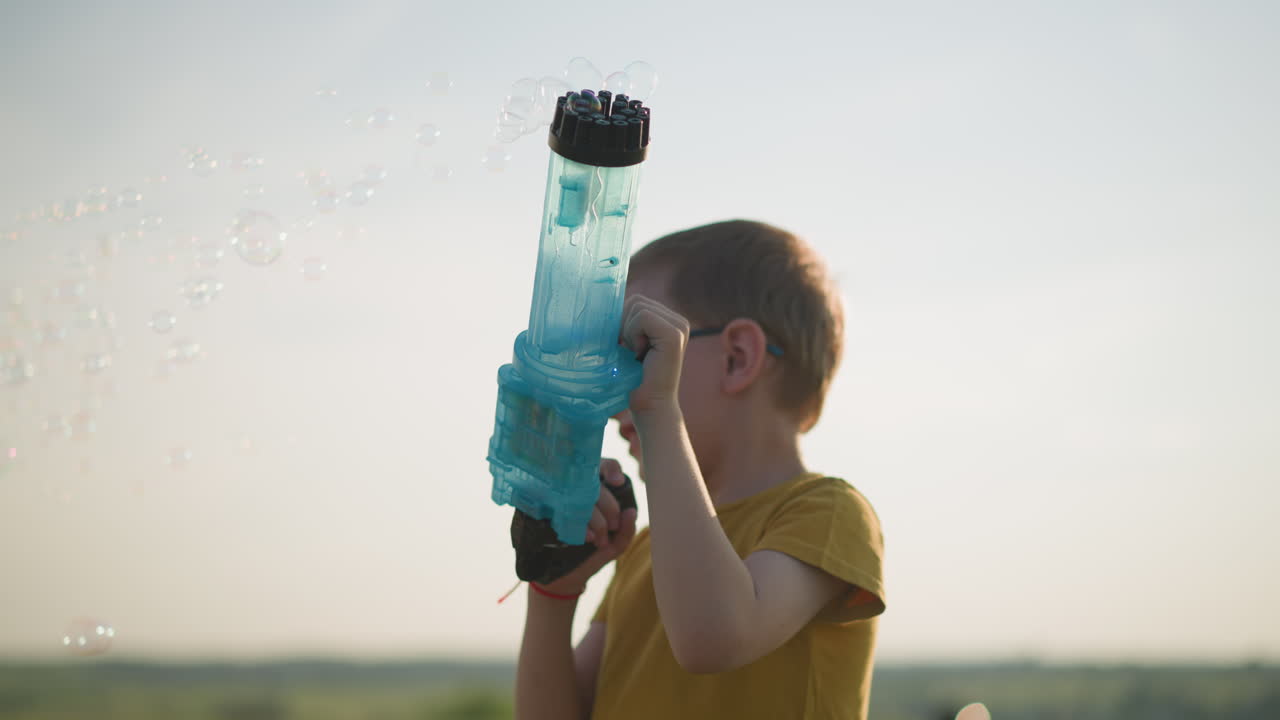 노란 셔츠와 안경을 입은 어린 소년은 야외에 서서 푸른 플라스틱 거품 총을 기게 들고 공중에 거품을 방출합니다.