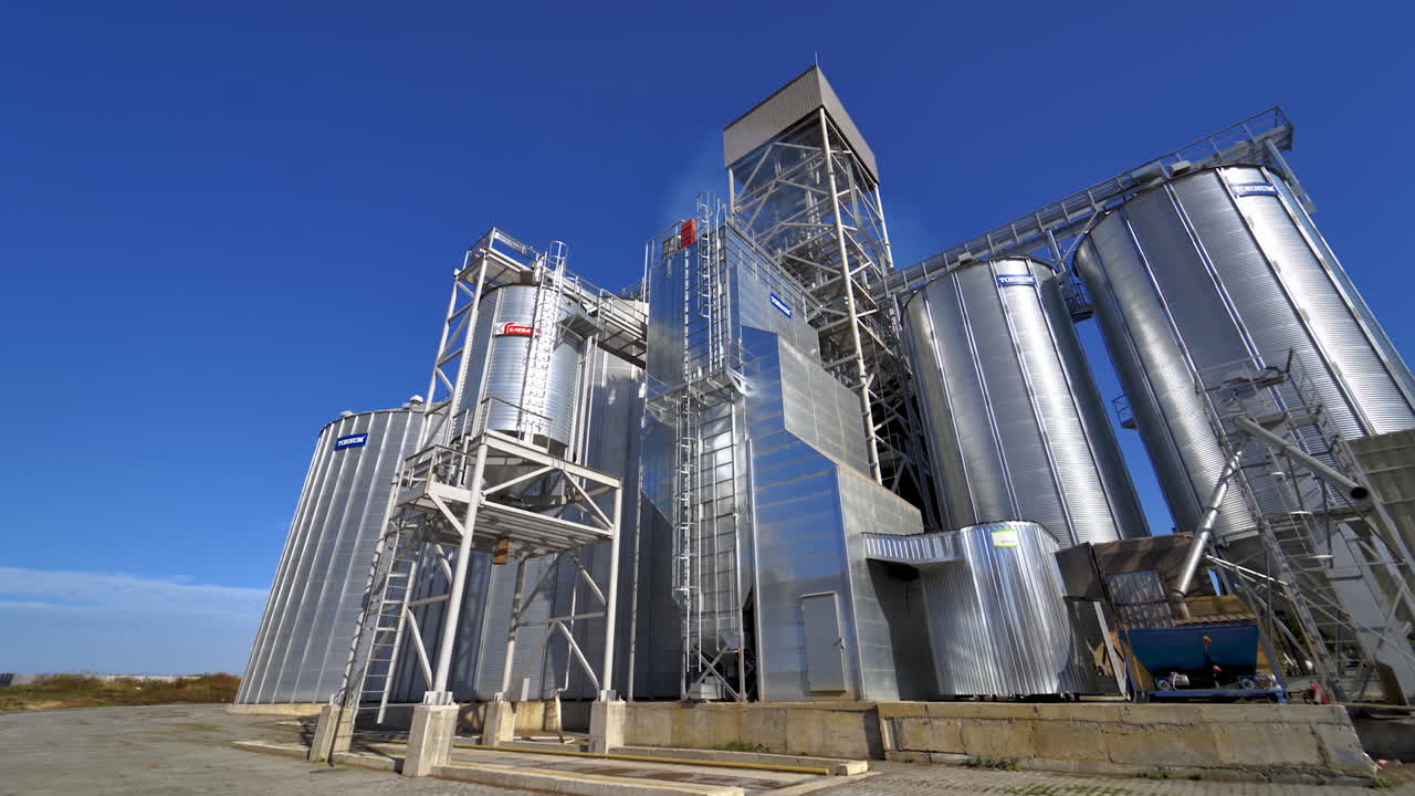 Modern plant for agribusiness. Silver grain elevators on blue sky background. Granary on field. Industrial factory with large storage bins at sunlight.