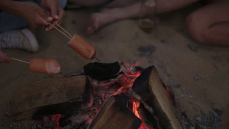 Nahaufnahme-Von-Würstchen,-Die-Am-Strandfeuer-Gegrillt-Werden.-Gruppe-Junger-Und-Fröhlicher-Menschen,-Die-Am-Feuer-Am-Strand-Sitzen