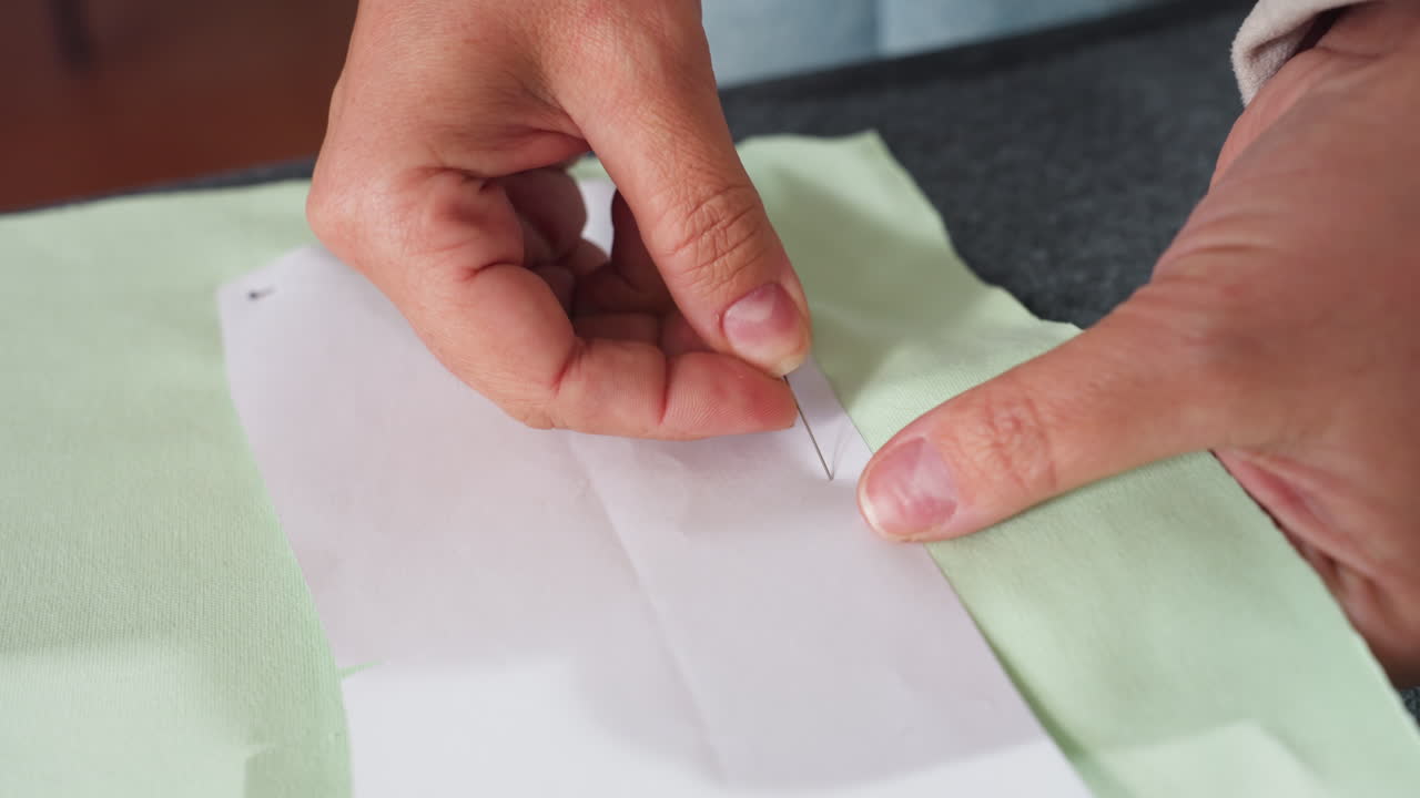 Close up of seamstress hand carefully securing white paper pattern onto light green textile using sharp needle in bright sewing studio preparing fabric for professional garment cutting process