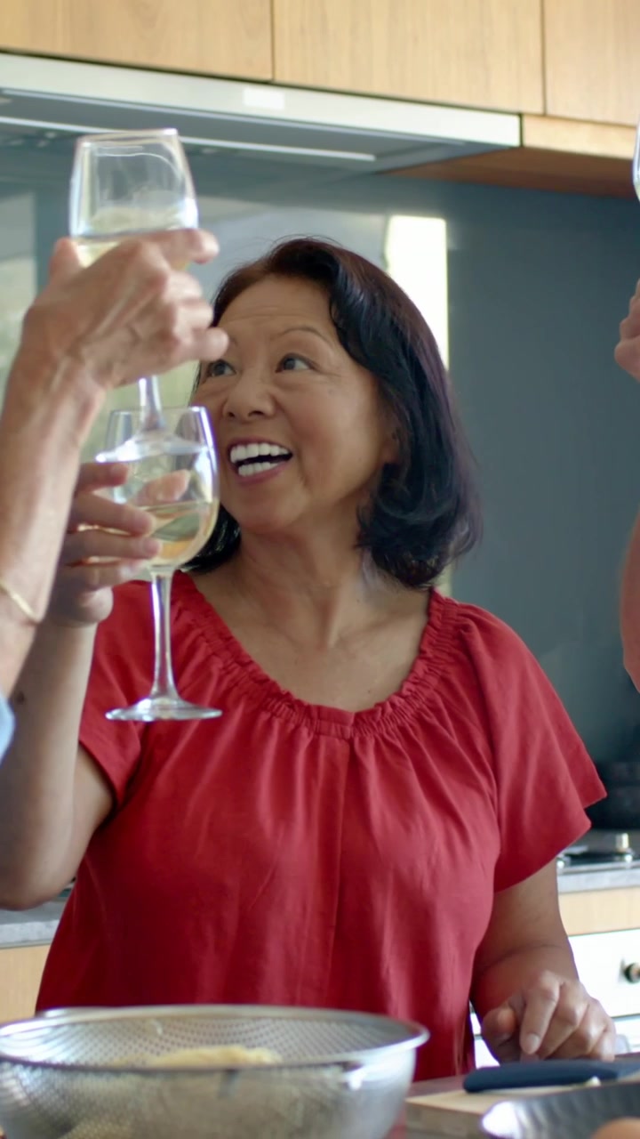 Vertical video: Clinking woman in red blouse raising wine glass in kitchen, friends joining toast