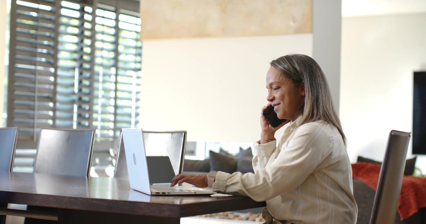 Donna afroamericana matura in camicia seduta a un tavolo, che usa telefono e laptop per lavoro, con spazio per il testo.