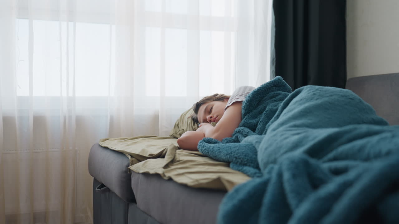 White Woman Napping On Sofa By Window With Blue Blanket, Sheer Curtains And Soft Morning Light Creating Peaceful Domestic Atmosphere, Close Side View, Relaxed Expression, Cozy Interior