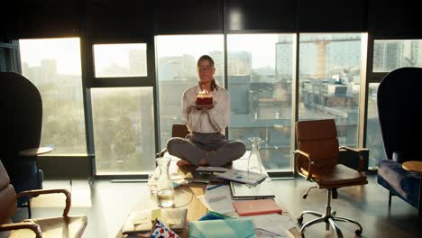 a girl in a white shirt sits on a table and blows candles on a cake in a sunny office with panoramic windows. happy celebration