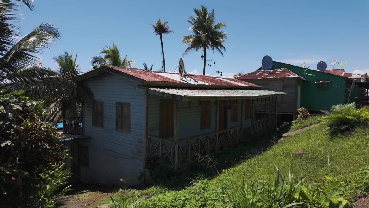 Casa sulla spiaggia con tetto in metallo arrugginito, antenne paraboliche in cima, circondata da lussureggianti piante tropicali e palme sotto un cielo azzurro e limpido in riva al mare.