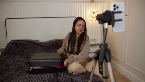 Happy-brunette-girl-in-a-brown-jacket-and-light-beige-things-in-a-black-suitcase-during-her-video-blog-and-preparing-to-go-on-vacation.-A-brunette-girl-collects-her-things-for-a-vacation-and-spends-time-for-her-own-pleasure.-Traveling