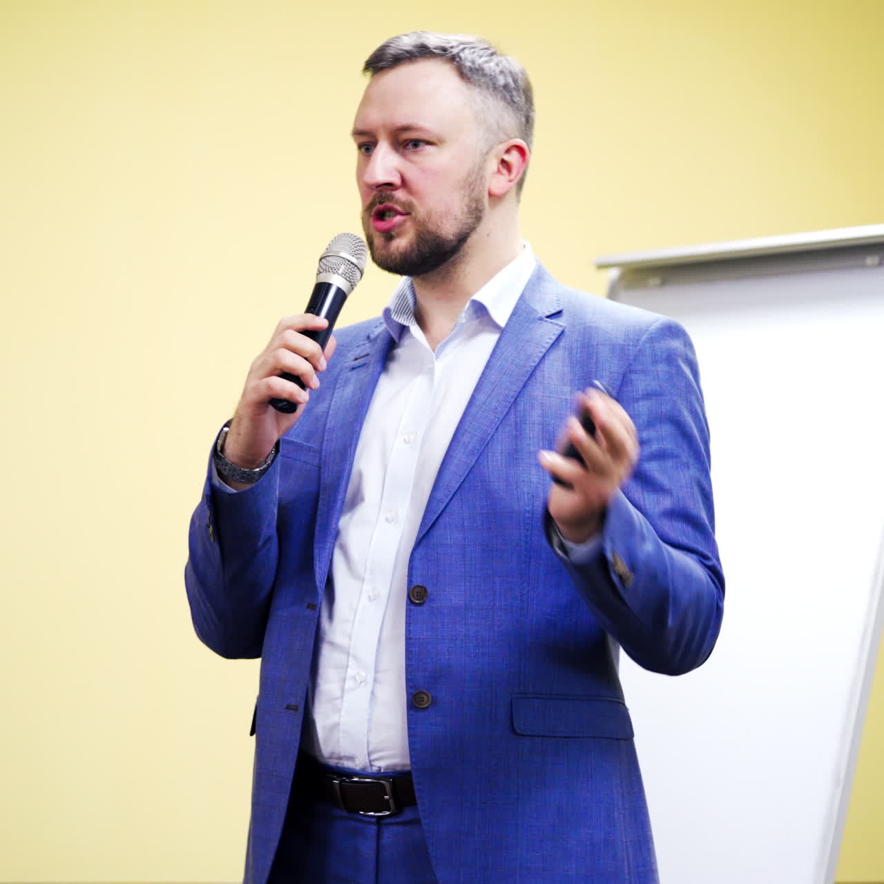 Bearded businessman in suit with microphone gives the lecture indoors. Professional speaker conducts speech gesturing a lot on business forum.