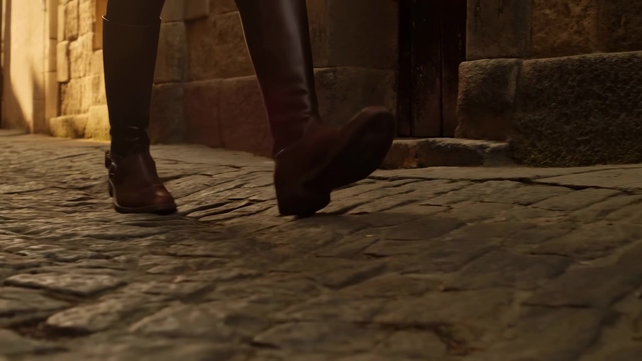 Primo piano ravvicinato a livello del suolo di gambe con pantaloni scuri e stivali di pelle alti al ginocchio, immortalate a metà passo su un vicolo acciottolato irregolare, accanto a un muro in muratura grezza.