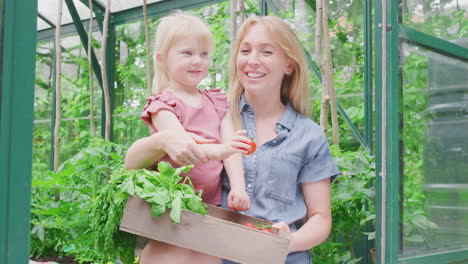 Mother-And-Daughter-Holding-Box-Of-Home-Grown-Vegetables-In-Greenhouse