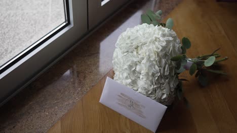 white floral bouquet with greenery and wedding invitation on wooden floor near window