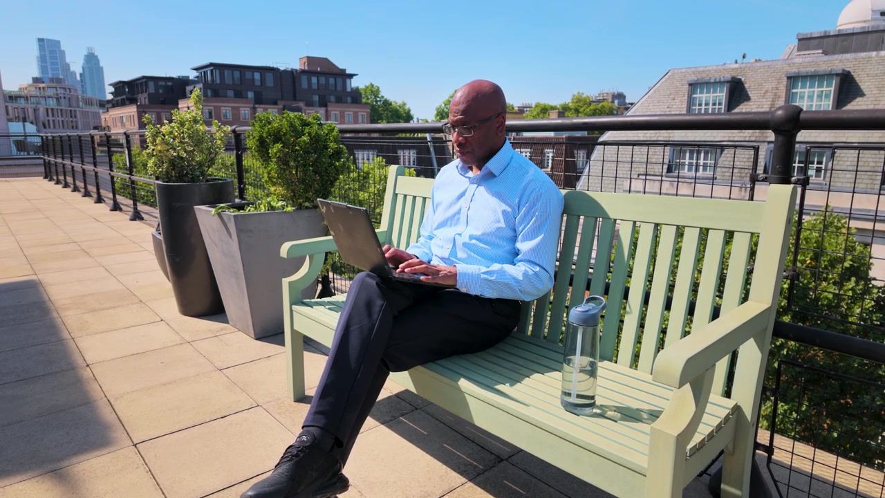 Imprenditore afroamericano che digita su un laptop e beve acqua, seduto su una panchina su una terrazza panoramica con vista sulla città sullo sfondo